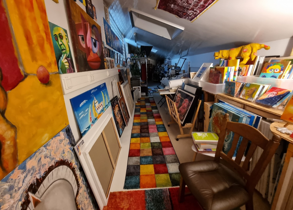 Foto meines Ateliers. Ein langer, gestreckter Raum, der links und rechts mit Kunst vollhängt und eine Menge Material zum Malen bietet.